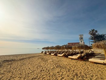 Οργανωμένη παραλία στην Αρτέμιδα με ψάθινες ομπρέλες και ξαπλώστρες, με φόντο τον γραφικό λευκό πύργο, ιδανική για χαλάρωση και καλοκαιρινό vibe.