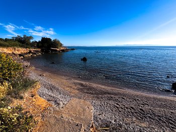 A tranquil, clear blue cove with a pebbly shore and rocky outcrops under a bright sky, characteristic of the beautiful beaches near Athens International Airport (ATH), such as Erotospilia or Vravrona, perfect for a quick swim or snorkeling during a layover.