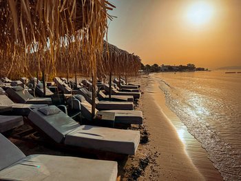 Golden hour view of an organized beach in Artemida (Loutsa), featuring rows of sunbeds and straw umbrellas along the calm, shallow waters, perfect for a relaxing day at a Blue Flag certified beach near Athens.