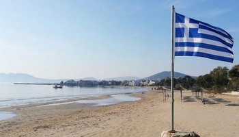 The Greek flag waving proudly over the beach in Artemida (Loutsa) under a clear blue sky, a symbol of March 25th celebrations.